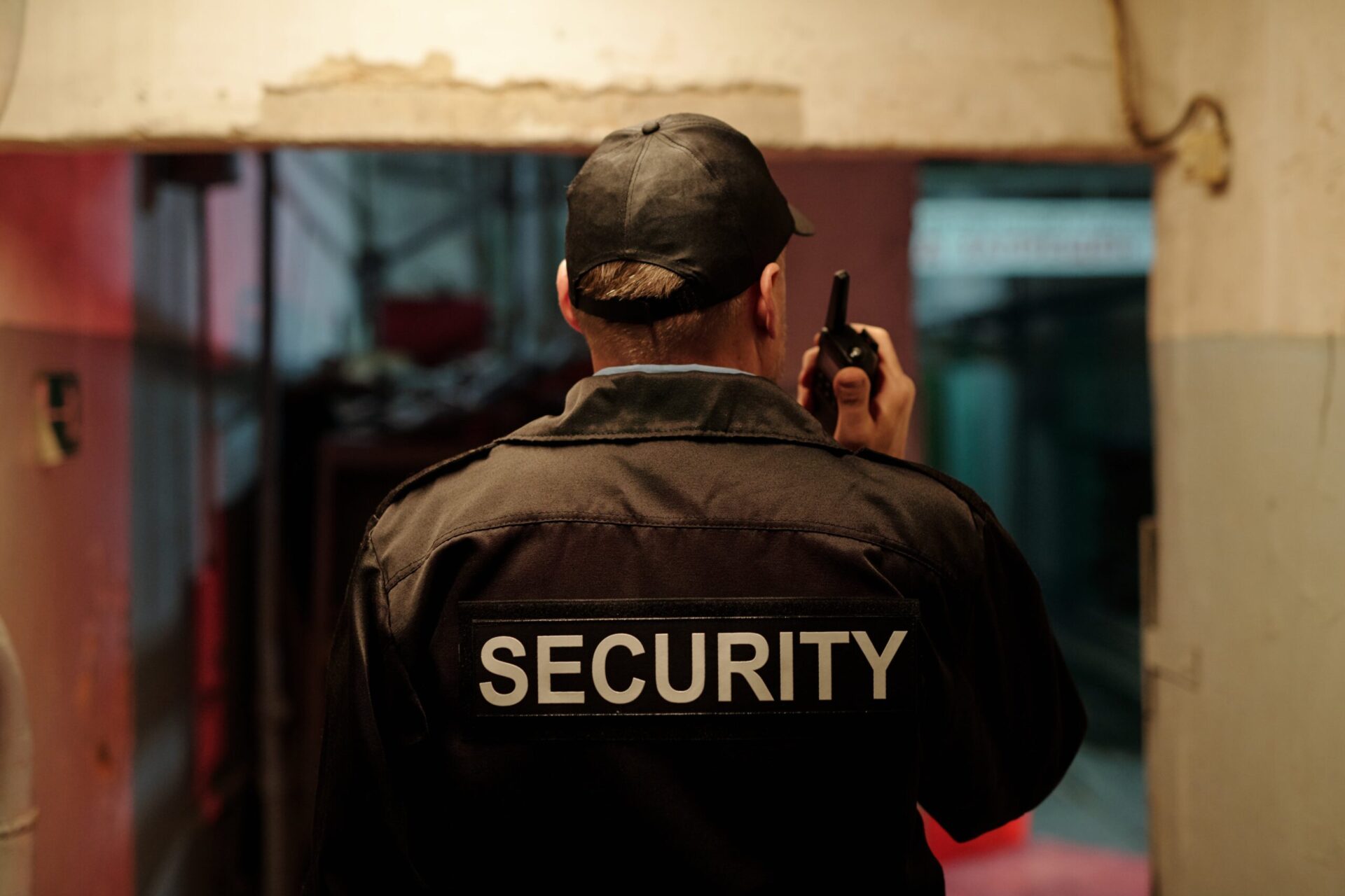 A security guard in a uniform and cap holding a walkie-talkie, facing away while patrolling an indoor area.